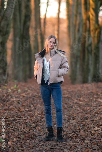 Young woman in a beige puffer jacket and blue jeans stands confidently on a leaf-covered path surrounded by tall trees in a serene forest setting