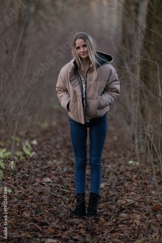 Young woman in a beige puffer jacket and blue jeans stands confidently on a leaf-covered path surrounded by tall trees in a serene forest setting