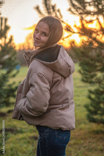Young woman in a beige puffer jacket and blue jeans stands confidently on a leaf-covered path surrounded by tall trees in a serene forest setting