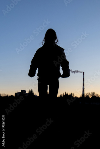 Young woman in a beige puffer jacket and blue jeans stands confidently on a leaf-covered path surrounded by tall trees in a serene forest setting