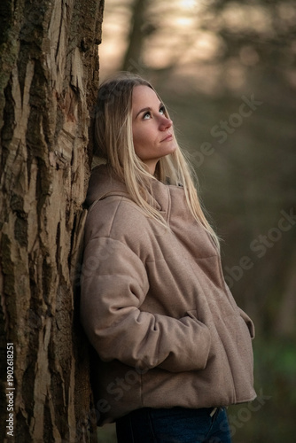 Young woman in a beige puffer jacket and blue jeans stands confidently on a leaf-covered path surrounded by tall trees in a serene forest setting