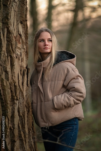 Young woman in a beige puffer jacket and blue jeans stands confidently on a leaf-covered path surrounded by tall trees in a serene forest setting