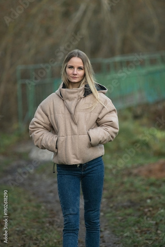 Young woman in a beige puffer jacket and blue jeans stands confidently on a leaf-covered path surrounded by tall trees in a serene forest setting