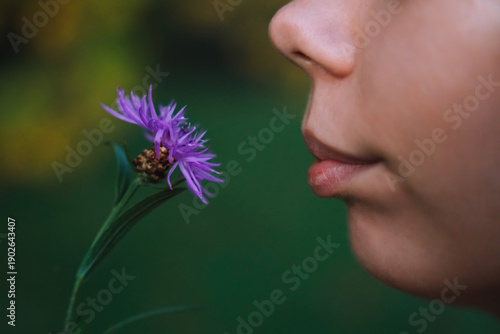 Side View Close Up Portrait of Young Woman Smelling Purple Wildflower Soft Lips Natural Skin Texture and Shallow Depth of Field in Green Outdoor Background