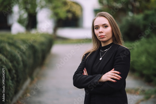 Confident Young Woman Standing with Crossed Arms in Park Wearing Black Jacket Natural Light Shallow Depth of Field and Soft Green Background