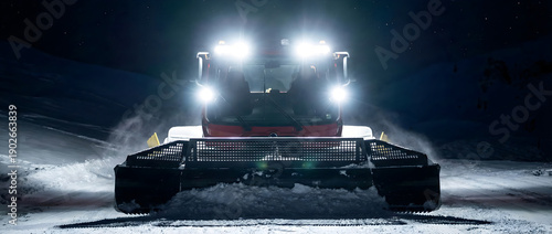 Snow groomer machine working on ski slope at night with bright headlights. Heavy snowcat vehicle preparing piste in ski resort.