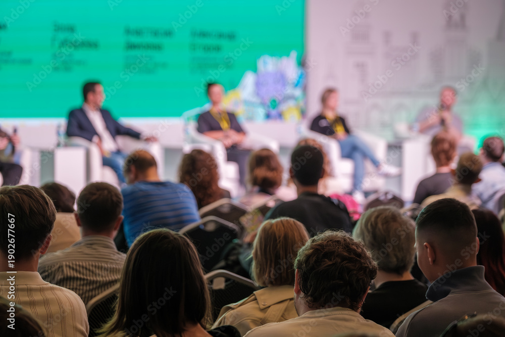 Obraz premium Panel discussion at conference with speakers on stage and attentive audience. Bright backdrop creates energetic mood.