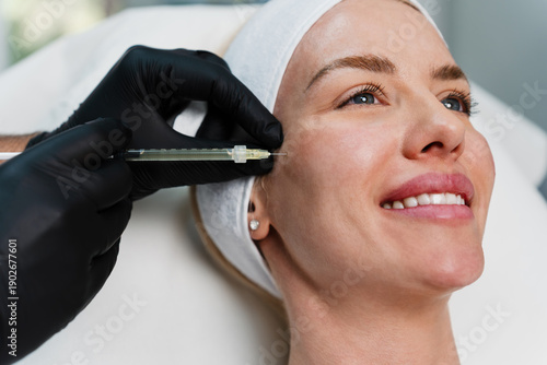 A smiling woman receives a professional facial filler injection in a modern aesthetic clinic. The procedure is performed by a doctor wearing black gloves.