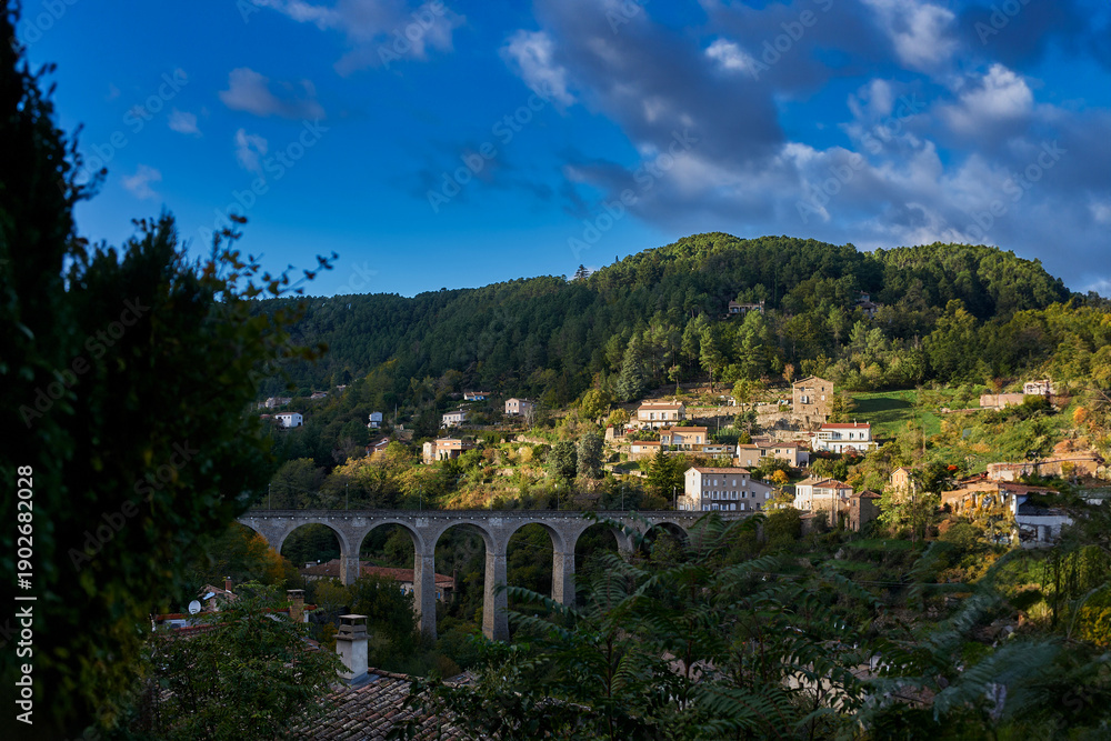 Fototapeta premium : Historic stone viaduct in the mountains near Privas, Ardeche. A majestic arched bridge surrounded by autumn-toned forests and traditional French countryside houses.