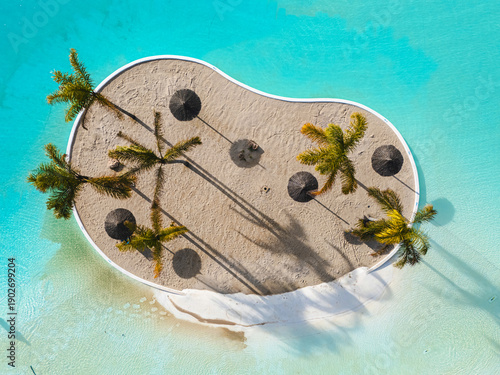 Aerial drone view artificial tropical lagoon in island beach surrounded by turquoise water, with palm trees and straw umbrellas. Vacation, summer holiday and relaxation