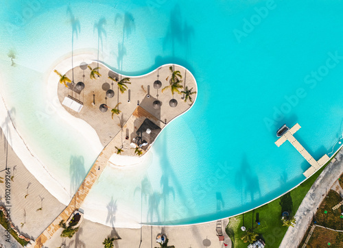 Aerial drone view artificial tropical lagoon in island beach surrounded by turquoise water, with palm trees and straw umbrellas. Vacation, summer holiday and relaxation