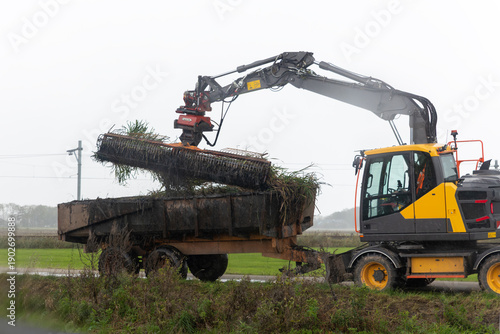 Fotografija Big modern excavator machine cutter arm cleaning overgrown grass reed ditch canal water at dutch countryside Netherlands village