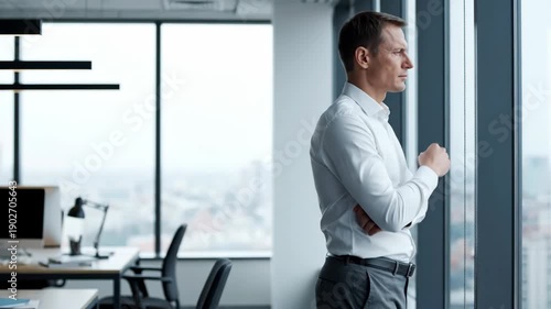 Professional man in office looking out window reflecting on city view. Corporate setting with modern workspace showcasing desk and chair. Concept for business meetings or corporate events.