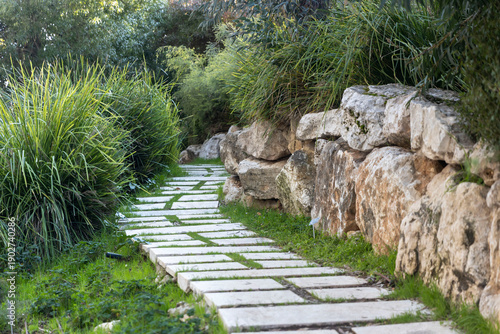 Stone paver pathway with grass growing between rectangular slabs, bordered by limestone rock wall and ornamental grasses in botanical garden setting.