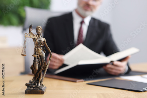 Notary in suit reading book at wooden table in office, focus on statue of Lady Justice