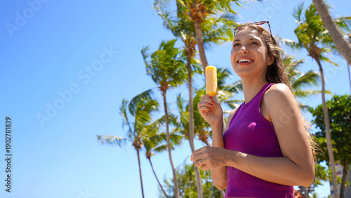 Happy young woman eating popsicle ice pop on the beach with palm trees on the background. Summer holidays concept.