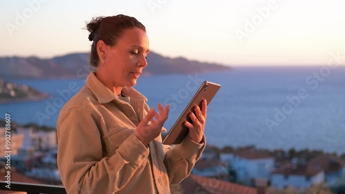 Woman on video call using tablet on balcony with sea view. Happy woman waving hand and talking on video call using her tablet on a balcony with a beautiful sea view during sunset, enjoying her travel