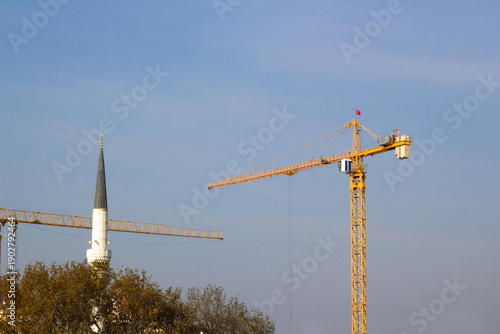 Foto Two yellow construction cranes against the blue sky, next to a white mosque minaret