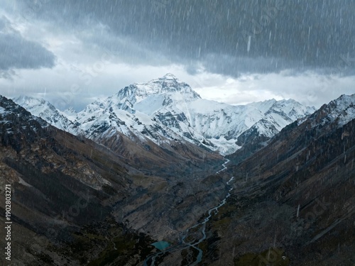 Snow-capped mountain range under stormy sky with rain falling over a rugged valley and river