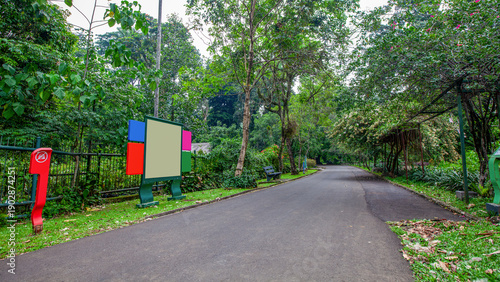 Winding asphalt pathway through a lush tropical botanical garden featuring modern, colorful blank information signage, dense foliage, and a blooming vine-covered arbor under soft daylight.
