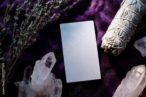 an empty, blank white tarot card on display, surrounded by crystals, dried herbs, and a sage smudge in the background, with a purple velvet cloth background