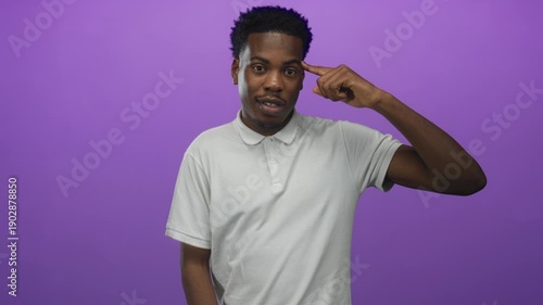 Young african american man in white polo shirt points finger to temple in studio against purple studio wall; playful madness.