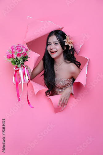 Lovely surprise for Valentine's: Elegant woman with a bouquet of pink roses breaking through pink paper wall.