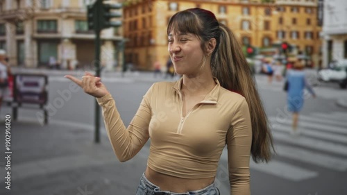 Woman points finger to street crosswalk by traffic lights and yellow building, visible midriff and smiling broadly; playful confidence.