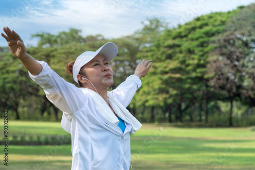 Middle-aged Asian woman exercises outdoors in a park. She raised her hand and took a deep breath of the fresh morning air, a smile on her face.