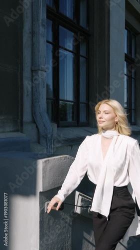 Stylish young woman walking along university terrace and leaning on balcony. Elegant female student or businesswoman enjoying city view on classic campus background.
