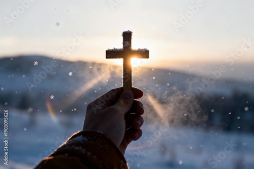 A hand holds a wooden cross against the backdrop of a sunset in winter. Snowflakes fall as the light shines through the cross. The scene shows a cold and serene setting.