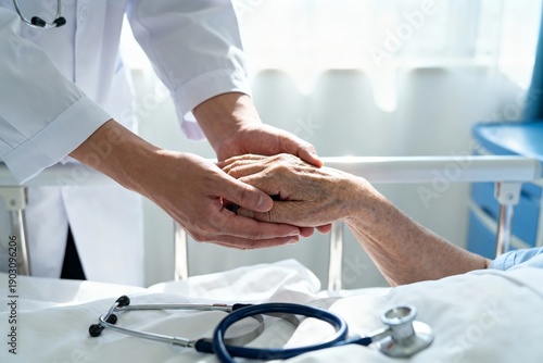 A healthcare professional holds the hand of an older patient in a hospital bed. The scene shows caring support during treatment. Sunlight comes through the window in the background.