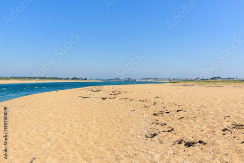Expansive sandy beach meets a vivid blue tidal channel at Barre d'Etel in Brittany. The scene is sunlit with clear sky, distant town, and natural coastal vegetation.