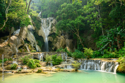 Golden sunlight illuminates the multi-level limestone cascades and turquoise pools of Kuang Si Waterfalls in northern Laos, surrounded by lush tropical forest. Vibrant green foliage and flowing water