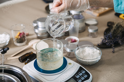 Photos Hand wearing glove pours clear liquid from beaker into glass jar on digital scale