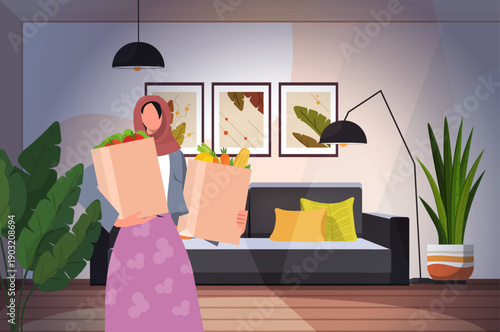 Plastic-free and zero waste sustainable lifestyle. Muslim woman with hijab standing in a modern living room, holding two grocery bags filled with fresh produce. Ethical consumerism. 