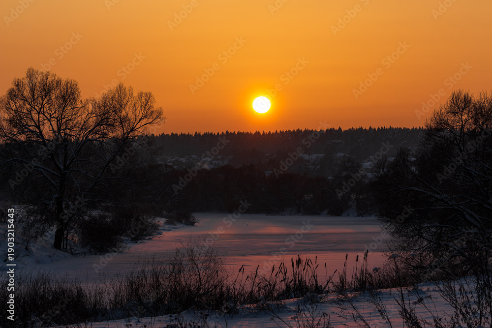Obraz premium Majestic winter sunset warms a tranquil frozen river, reflecting golden hues on crisp snow. Bare trees silhouette against a brilliant sky, with a hint of distant dwellings