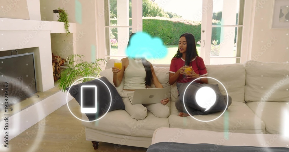 Fototapeta premium Sharing two women sipping orange juice and using silver laptop on sofa in bright living room