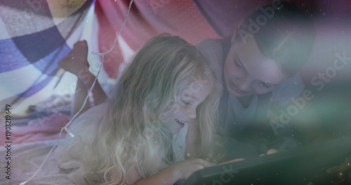Lying two girls watching tablet in DIY blanket fort, wearing pajamas, with pillows and fairy lights