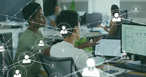 Pointing Korean colleague guiding peer at monitors in open office in business-casual, showing UI