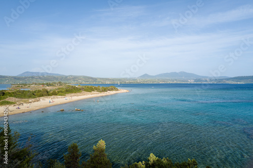 Fotografie Expansive view of a sandy beach curving along the transparent blue water, with scattered sunbathers and lush greenery, set against distant hills on the Methoni coast in Messenia, Peloponnese
