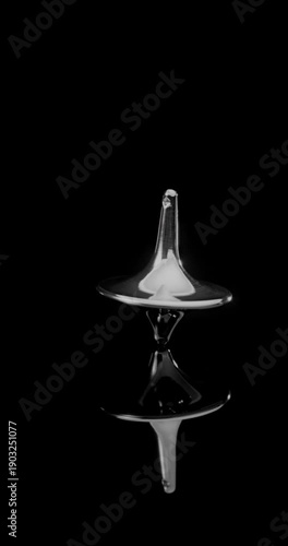 Metallic totem in initial steady rotation, gradually increasing wobble and tilt toward collapse against a deep black mirrored backdrop.