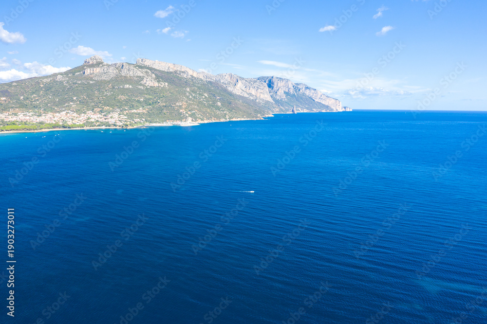 Fototapeta premium Wide aerial view of the deep blue Mediterranean Sea meeting the rugged green mountains and coastline near Lotzorai, Sardinia. The scene is bright and tranquil with a clear sky and subtle waves.