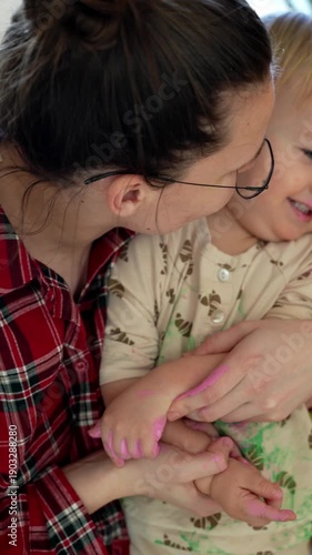 mother hugging a toddler while sitting near a decorated christmas tree indoors. adult holding the child from behind. holiday home setting with soft natural light.
