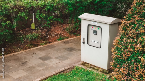 Industrial electrical panel next to sidewalk with lush green bush and grass wallpaper background