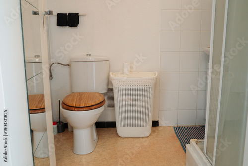 White bathroom interior with a toilet featuring a wooden seat cover and a white laundry hamper. Clean everyday home scene focused on simple bathroom furniture and neutral interior details.