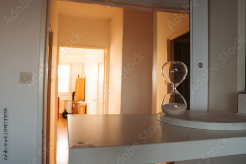 Hourglass on a white shelf with a sunlit apartment corridor in the background. Simple interior scene with natural light and everyday home objects.
