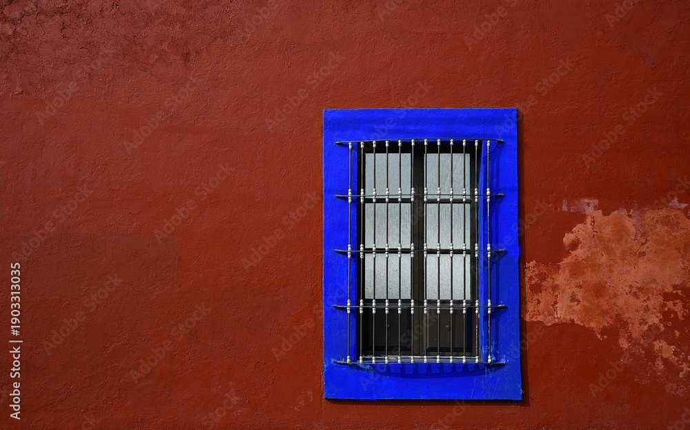 Obraz premium Old traditional Colonial house window with handcrafted wrought iron grilles and an electric blue trim on a Venetian red stucco wall in Cholula, Puebla Mexico. 