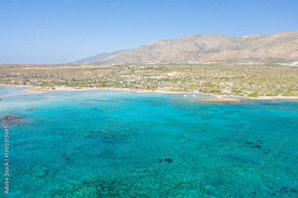 Obraz premium Expansive aerial perspective of Elafonisi beach in Crete, showcasing clear turquoise sea, sandy shoreline, and rugged hills beneath a cloudless blue sky. The vivid colors and open landscape evoke a