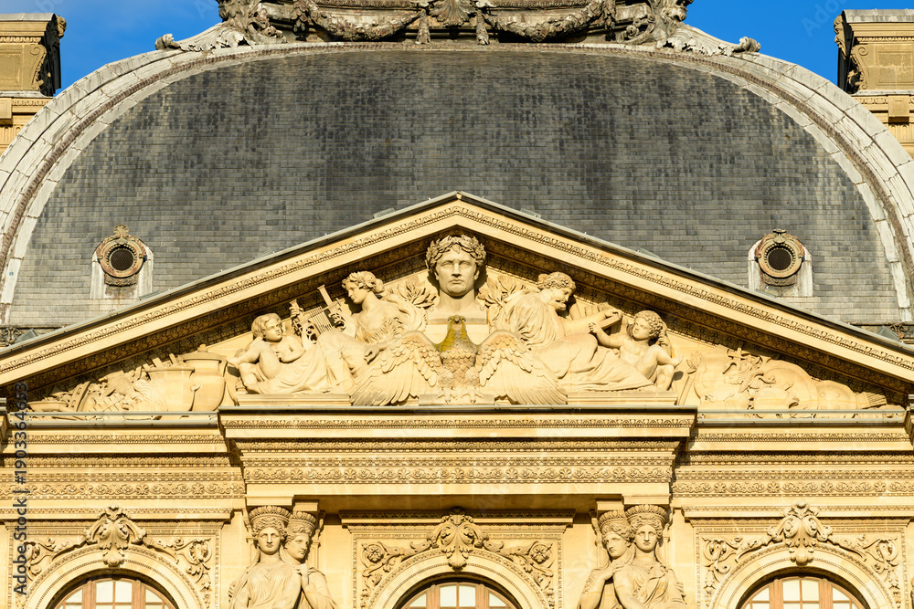 Obraz premium Close view of the ornate pediment on the Louvre palace in Paris, France, featuring detailed classical stone sculptures illuminated by warm sunlight. The historic reliefs and slate dome create a